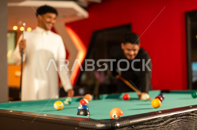 الانشطة الشبابية الممتعة داخل المملكة , رجل عربي خليجي سعودي يرتدي الثوب الاسود يحمل العصا الخشبية الخاصة بالبلياردو , القيام بتحريك الكرات الصغيرة الملونة, مشاركة الاصدقاء الاوقات الممتعة