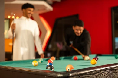 Fun youth activities inside the kingdom, an Arab Gulf Saudi man wearing a black thobe holding a wooden cue for billiards, moving the small colored balls, sharing enjoyable times with friends.