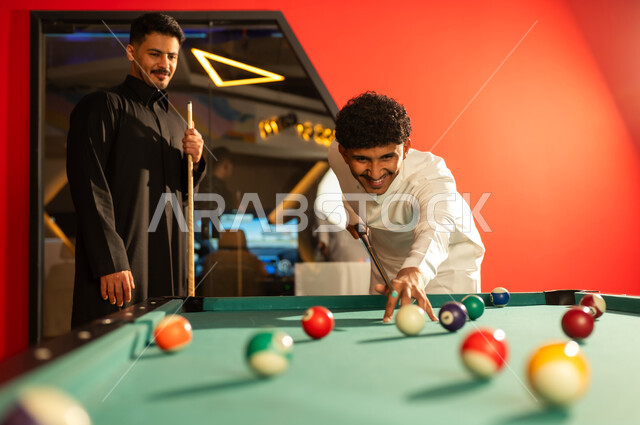 رجل عربي خليجي سعودي يرتدي الثوب الابيض يقوم بتحريك الكرة البيضاء بالعصا الخشبية الخاصة بلعبة البلياردو , ضربة البداية اشارة لبدء اللعب , صديقه يرتدي الثوب الاسود مستنداً على الطاولة ينتظر دوره في اللعب