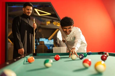 رجل عربي خليجي سعودي يرتدي الثوب الابيض يقوم بتحريك الكرة البيضاء بالعصا الخشبية الخاصة بلعبة البلياردو , ضربة البداية اشارة لبدء اللعب , صديقه يرتدي الثوب الاسود مستنداً على الطاولة ينتظر دوره في اللعب