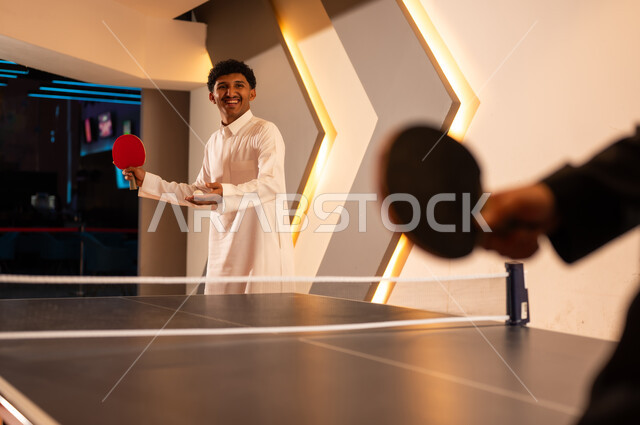 He holds the small white ball for table tennis in the palm of his hand, a Saudi Arabian man wearing a white thobe smiles at his friend, fun moments with friends.