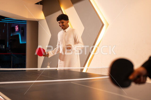 النظر الى ارتطام الكرة الصغيرة بالطاولة , رجل عربي خليجي سعودي يرتدي الثوب الابيض يتابع الكرة الصغيرة للفوز على المنافس , اللعب مع الاصدقاء بسعادة و فرحة