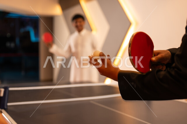 A close-up image of a small white table tennis ball, two Arab Gulf Saudi men wearing a white thobe and a black thobe are having a good time inside gaming centers, using a small red paddle to make the opening shot.