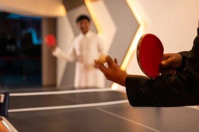 صورة مقربة لكرة تنس طاولة بيضاء صغيرة , رجلان عربيان خليجيان سعوديان يرتديان الثوب الابيض و الثوب الاسود يقضيان اوقات مرحة داخل مراكز الالعاب , استخدام المضرب الاحمر الصغير للقيام بضربة البداية