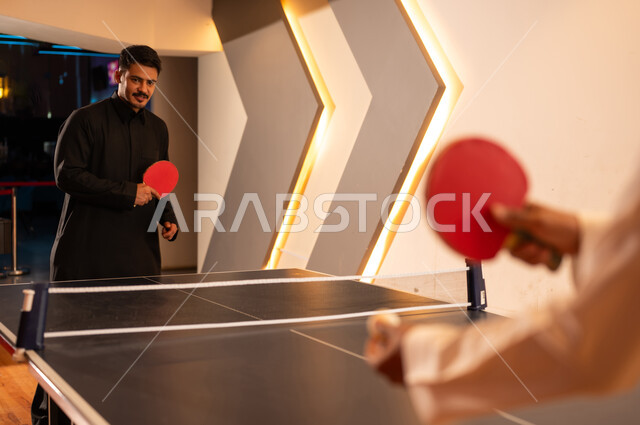 متابعة الكرة بتركيز , رجل عربي خليجي سعودي يرتدي الثوب الاسود يحمل المضرب الخشبي الاحمر الصغير , الروح التنافسية بين الاصدقاء , شاب عربي خليجي سعودي يرتدي الثوب الابيض يحمل الكرة البيضاء