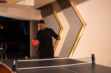 ضربة البداية بلعبة تنس الطاولة , رجل عربي خليجي سعودي يرتدي الثوب الاسود يضرب الكرة الصغيرة بالمضرب الاحمر , لحظات ممتعة و ملسية مع صديقه , انشطة شبابية داخل مراكز الالعاب بالمملكة