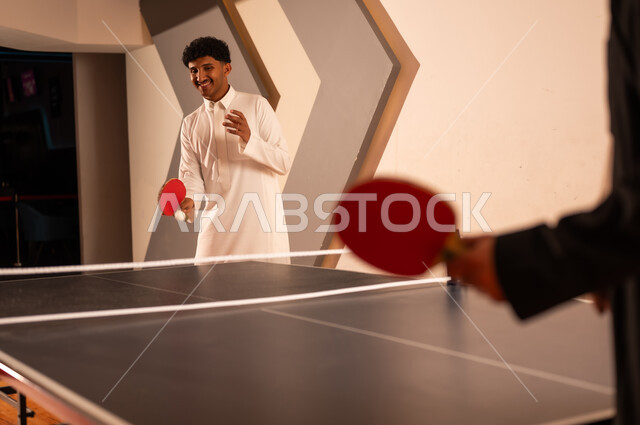 رجل عربي خليجي سعودي يرتدي الثوب الابيض ينظر الى ارتطام الكرة بالمضرب الاحمر الصغير , طاولة تنس طاولة منقسمة لنصفين بالشبكة , اوقات سعيدة ممتعة مع الاصدقاء