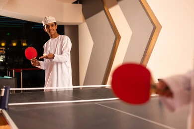 Looking happily at the camera, a young Arab Gulf Omani man wearing a white dishdasha and a embroidered kumma stands in front of a small table tennis table, holding a small white ball in the palm of his hand, with gestures of happiness.