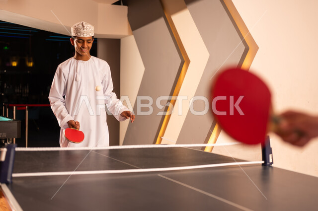 متابعة الكرة الصغيرة في الهواء بعد رميها , شاب عربي خليجي عماني يرتدي الدشداشة البيضاء و الكمة المطرزة يحمل المضرب الصغير الخاص بلعبة تنس الطاولة , ايماءات السعادة و الابتسامة