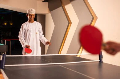 متابعة الكرة الصغيرة في الهواء بعد رميها , شاب عربي خليجي عماني يرتدي الدشداشة البيضاء و الكمة المطرزة يحمل المضرب الصغير الخاص بلعبة تنس الطاولة , ايماءات السعادة و الابتسامة