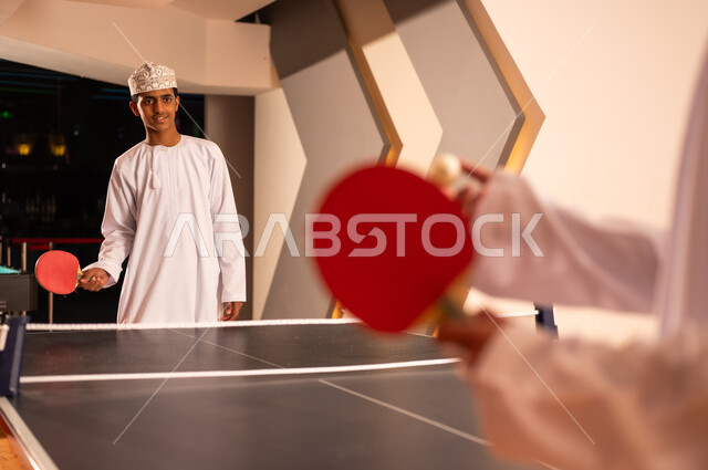 الالعاب الشبابية الجماعية الممتعة داخل سلطنة عمان , صديقان عربيان خليجيان عمانيان يرتديان دشداشة بيضاء و كمة مطرزة يلعبون تنس الطاولة , ايماءات الفرحة و مشاركة اللحظات السعيدة