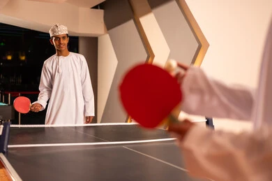The enjoyable collective youth games inside the Sultanate of Oman, two Arab Gulf friends from Oman wearing white dishdasha and embroidered kumma are playing table tennis, gestures of joy and sharing happy moments.