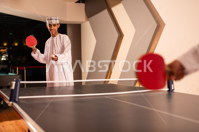 النظر للكرة الصغيرة قبل بداية اللعبة , شاب عربي خليجي عماني يرتدي دشداشة بيضاء و كمة مطرزة يحمل مضرب احمر صغير , طاولة تنس خشبية مقسمة نصفين بشبكة , لعبة جماعية