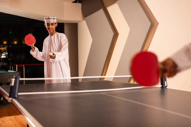 Looking at the small ball before the game starts, a young Arab Gulf Omani man wearing a white dishdasha and an embroidered kumma holds a small red racket, a wooden table tennis table divided in half by a net, a team game.
