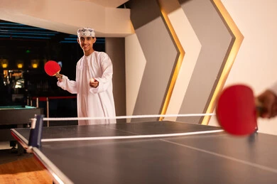 رمي كرة تنس الطاولة الصغيرة للاشارة لبداية اللعبة المسلية , شاب عربي خليجي عماني يرتدي الدشداشة البيضاء و الكمة المطرزة يحمل المضرب الاحمر الصغير , الالعاب الممتعة الجماعية بين الاصدقاء