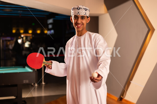 شاب عربي خليجي عماني يرتدي الدشداشة البيضاء و الكمة المطرزة يحمل المضرب الاحمر الصغير , النظر و الابتسامة الي الكاميرا , صورة مقربة , كرة صغيرة بيضاء خاصة بلعبة تنس الطاولة