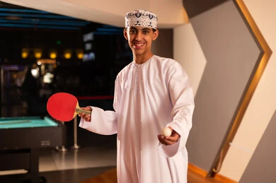 A young Arab Gulf Omani man wearing a white dishdasha and a embroidered kumma holds a small red paddle, looking and smiling at the camera, a close-up shot, a small white ball specifically for table tennis.