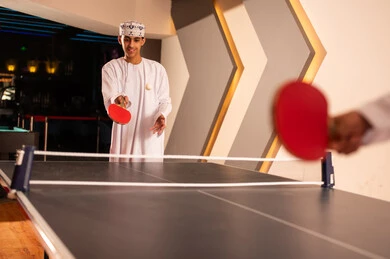 طاولة خاصة بلعبة تنس الطاولة مقسمة نصفين بشبكة , شاب عربي خليجي عماني يرتدي الدشداشة البيضاء و الكمة المطرزة يحمل المضرب الاحمر الصغير , رمى الكرة للاشارة الي بداية اللعبة , ايماءات السعادة و الابتسامة اثناء قضاء الاوقات الممتعة طاولة خاصة بلعبة تنس الطاولة مقسمة نصفين بشبكة , شاب عربي خليجي عماني يرتدي الدشداشة البيضاء و الكمة المطرزة يحمل المضرب الاحمر الصغير , رمى الكرة للاشارة الي بداية اللعبة , ايماءات السعادة و الابتسامة اثناء قضاء الاوقات الممتعة