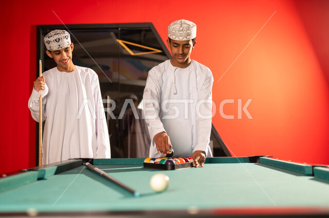 شاب عربي خليجي عماني يرتدي الدشداشة البيضاء و الكمة المطرز يضع الكرة السوداء في المثلث بعد نهاية اللعبة , الوقوف امام طاولة البلياردو الخشبية , ايماءات السعادة والابتسامة لقضاء الوقت الممتع مع الاصدقاء