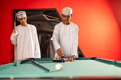 A young Arab Gulf Omani man wearing a white dishdasha and a embroidered kumma places the black ball in the triangle after the end of the game, standing in front of the wooden billiard table, gestures of happiness and smiles enjoying quality time with friends.