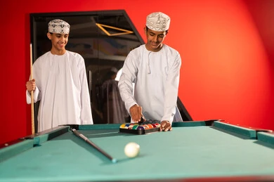 النظر الى الكرات الصغيرة الملونة المرتبة داخل المثلث الاسود, شابان عربيان خليجيان عمانيان يرتديان الدشداشة البيضاء و الكمة المطرزة يقفان امام طاولة البلياردو , اوقات سعيدة مسلية بين الاصدقاء