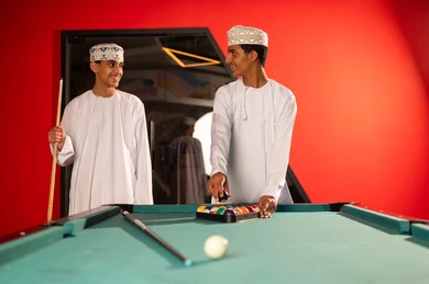 Two young Arab Gulf Omanis wearing white dishdasha and embroidered kumma stand in front of the green billiard table, placing the black ball in the triangle, arranging the small colored balls, finishing the game, gestures of happiness and smiles at each other.