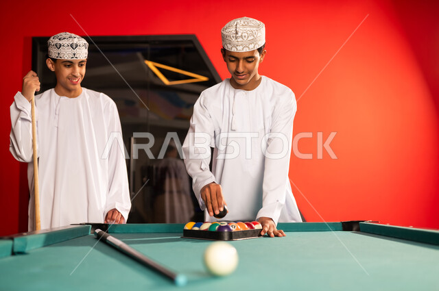 شاب عرب خليجي عماني يرتدي الدشداشة البيضاء والكمة المطرزة يضع الكرة السوداء داخل المثلث الاسود , ترتيب الكرات الصغيرة بعد الانتهاء من اللعب , مفهوم الحفاظ على نظافة المكان , ايماءات السعادة و الابتسامة للكاميرا