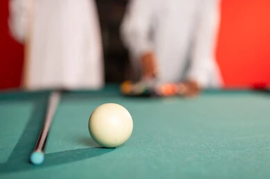 A close-up image of a white billiard ball, two Arab Gulf young men from Oman wearing white dishdasha standing in front of a billiard table, the wooden cue for the game on the green table, the small balls arranged inside the black triangle.