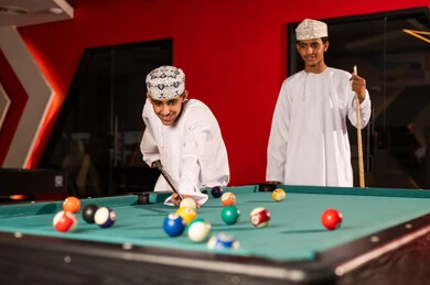 الانشطة الترفيهية الشبابية بسلطنة عمان , شابان عربيان خليجيان عمانيان يرتديان الدشداشة البيضاء و الكمة المطرزة يقفان امام طاولة البلياردو , استخدام العصا الخشبية لتحريك الكرات الصغيرة الملونة