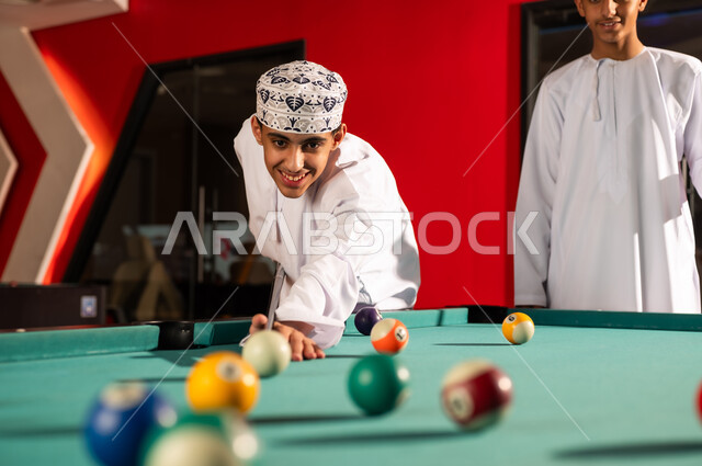 رجل عربي خليج عماني يرتدي الدداشة البيضاء و الكمة المطرزة يحرك الكرة البيضاء الصغيرة بالعصا الخشبية , لحظات ممتعة بين الاصدقاء داخل مركز الالعاب , النظر الي الكرات الصغيرة الملونة