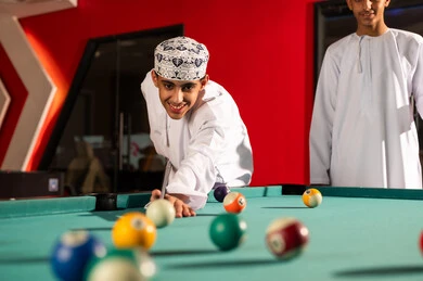 An Omani Gulf Arab man wearing a white dishdasha and an embroidered kumma moves a small white ball with a wooden stick, enjoying moments with friends inside the game center, looking at the small colored balls.