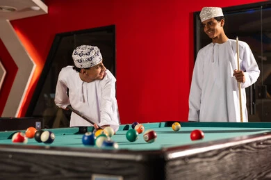قضاء الاوقات المسلية بين الاصدقاء , شابان عربيان خليجيان عمانيان يرتديان دشداشة بيضاء و كمة مطرزة يقفان امام طاولة البلياردو , تحريك الكرات الملونة الصغيرة باستخدام العصا الخشبية , ايماءات السعادة