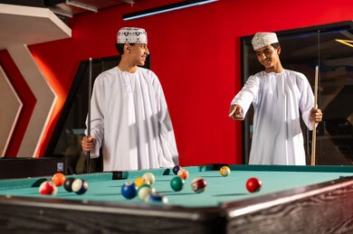 A young Arab Gulf Omani man wearing a white dishdasha and an embroidered kumma is looking at his friend with a smile, enjoying fun youth activities inside gaming centers, hitting small balls with the wooden stick used for billiards.