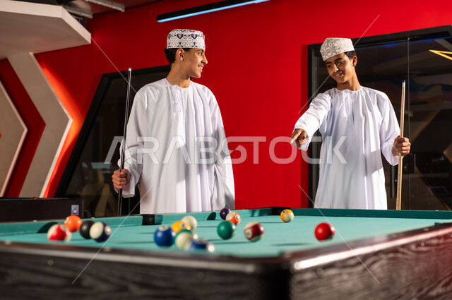 Standing in front of the billiard table, two young Arab Gulf Omanis wearing white dishdashas and embroidered kumas hold the wooden cues specific to the game, pointing with their index fingers at the small balls, smiling and happy while playing.