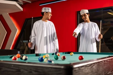 الوقوف امام طاولة البلياردو , شابان عربيان خليجيان عمانيان يرتديان دشداشة بيضاء و كمة مطرزة يحملان العصيان الخشبية الخاصة باللعبة , الاشارة باصبع السبابة الي الكرات الصغيرة , الابتسامة و السعادة اثناء اللعب
