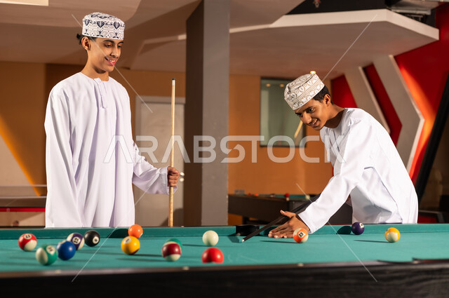 Preparing to start a game of billiards, two young Arab Gulf Omanis wearing embroidered kumma and white dishdasha, standing in front of the game table, small colored numbered balls, happy gestures enjoying fun times with friends.