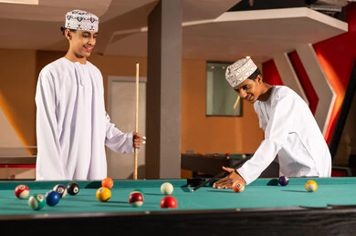 الاستعداد لبداية لعبة البلياردو , شابان عربيان خليجان عمانيان يرتديان كمة مطرزة و دشداشة بيضاء , الوقوف امام طاولة اللعبة , كرات ملونة صغيرة مرقمة , ايماءات سعيدة بقضاء الاوقات المسلية مع الاصدقاء