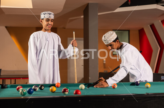 النظر الى كرات البلياردو الصغيرة الملونة , شابان عربيان خليجيان عمانيان يرتديان الدشداشة البيضاء و الكمة المطرزة يقفان امام الطاولة , الاستعداد و الحماس اثناء اللعب مع الاصدقاء