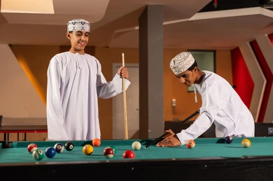 Looking at the small colored billiard balls, two young Arab Gulf Omanis wearing white dishdasha and embroidered kumma stand in front of the table, ready and excited while playing with friends.