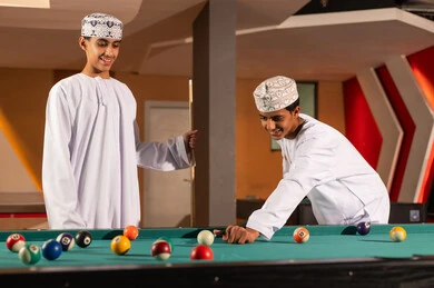 Two Arab Gulf young men from Oman wearing white dishdasha and embroidered kumma are holding the wooden cue stick for the billiards game, striking the small colored balls, playing with high focus and professionalism, gestures of happiness and smiles, close-up image.