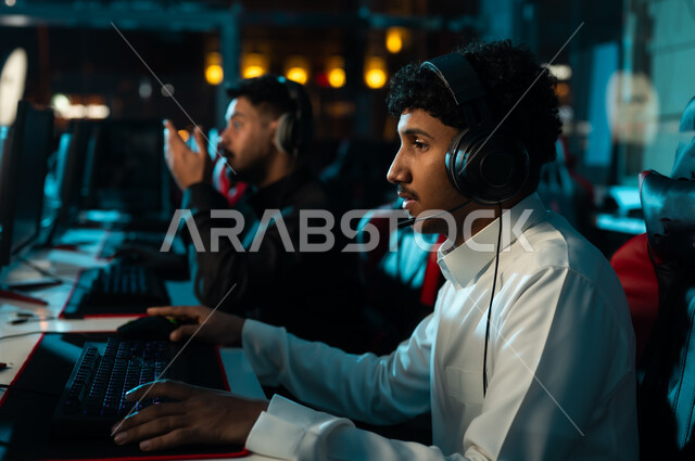 شاب عربي خليجي سعودي يرتدي الثوب الابيض يجلس بتركيز و شغف امام شاشة الحاسب المكتبي , ألعاب الكترونية ممتعة , وضع سماعة الرأس للتواصل المباشر مع الاصدقاء اثناء اللعب , صورة مقربة , رجل عربي خليجي سعودي يرتدي الثوب الاسود
