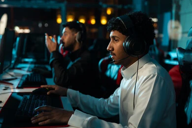 شاب عربي خليجي سعودي يرتدي الثوب الابيض يجلس بتركيز و شغف امام شاشة الحاسب المكتبي , ألعاب الكترونية ممتعة , وضع سماعة الرأس للتواصل المباشر مع الاصدقاء اثناء اللعب , صورة مقربة , رجل عربي خليجي سعودي يرتدي الثوب الاسود