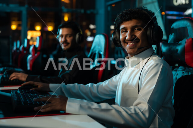 النظر الي الكاميرا بسعادة و ابتسامة , رجلان عربيان خليجيان سعوديان يرتديان الثوب الابيض و الاسود يقضيان اوقات مسلية في مركز الالعاب الالكترونية , العاب جماعية شبابية داخل المملكة
