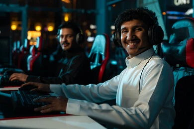 النظر الي الكاميرا بسعادة و ابتسامة , رجلان عربيان خليجيان سعوديان يرتديان الثوب الابيض و الاسود يقضيان اوقات مسلية في مركز الالعاب الالكترونية , العاب جماعية شبابية داخل المملكة