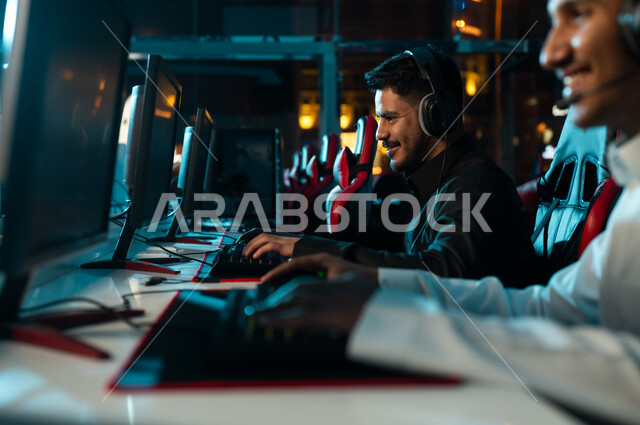 رجل عربي خليجي سعودي يرتدي الثوب الاسود يجلس بجانب صديقه , اللعب و التركيز الشديد في شاشة الحاسب المكتبي , العاب الكترونية ممتعة , اوقات جماعية مسلية بين الاصدقاء