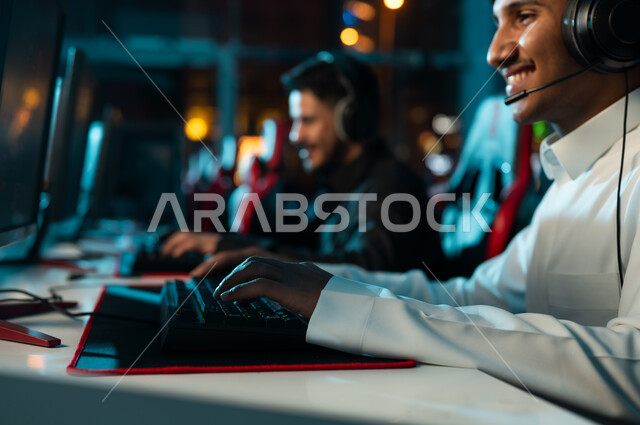 رجل عربي خليجي سعودي يرتدي الثوب الابيض يجلس بجانب صديقه بالثوب الاسود , اللعب مع الاصدقاء عن بعد باستخدام الانترنت , انشطة جماعية ممتعة في مراكز الالعاب الالكترونى , صورة مقربة