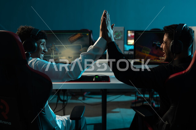 Celebrating the victory by high-fiving each other, two Arab Gulf men from Saudi Arabia, one wearing a white thobe and the other a black thobe, are sitting in an electronic gaming center, with expressions of overwhelming joy and big smiles.
