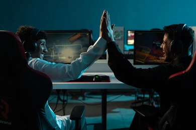 Celebrating the victory by high-fiving each other, two Arab Gulf men from Saudi Arabia, one wearing a white thobe and the other a black thobe, are sitting in an electronic gaming center, with expressions of overwhelming joy and big smiles.
