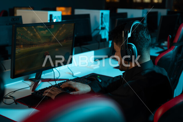مركز الألعاب الالكترونية يحتوي على اجهزة تقنية متعددة , رجل عربي خليجي سعودي يرتدي الثوب الاسود يجلس امام شاشة الحاسب , اللعب بتركيز و حماس , انشطة ترفيهية شبابية بالمملكة