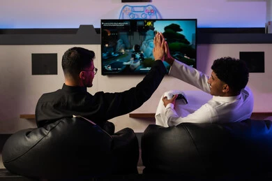 Clapping hands together is a sign of victory and reaching the final level in the video game. Two Arab Gulf men from Saudi Arabia, wearing a white thobe and a black thobe, are sitting on comfortable sponge seats, showing gestures of joy and enjoying happy times with friends.